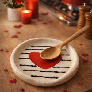 May include: A ceramic plate with a red heart design and a wooden spoon. The plate has black stripes and is surrounded by red heart-shaped confetti. Red candles and roses are in the background.