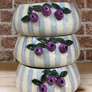 May include: Three stacked ceramic bowls with a cream and blue striped pattern. Each bowl is decorated with three purple blueberry-like embellishments and green leaves. The bowls are stacked on top of each other.