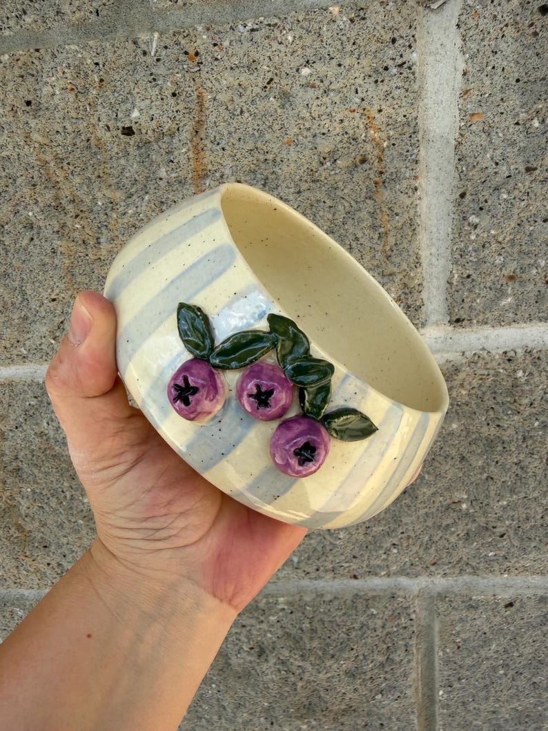 May include: A ceramic bowl with a cream-colored interior and exterior. The exterior features blue and cream stripes and three purple blueberry clusters with green leaves. The bowl is held in a hand.