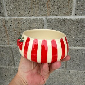 May include: A ceramic bowl with a cream-colored interior and exterior. The exterior features vertical red stripes alternating with cream-colored stripes. A cluster of red strawberries with green leaves decorates the side of the bowl.