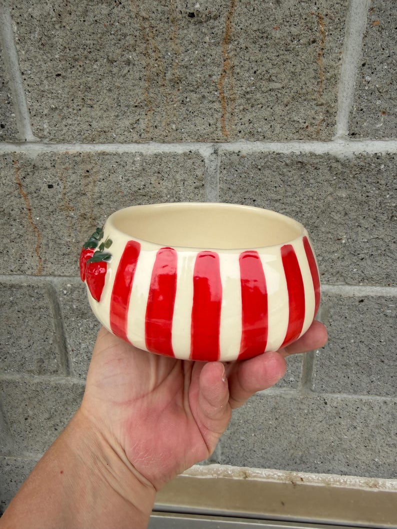 May include: A ceramic bowl with a cream-colored interior and exterior. The exterior features vertical red stripes alternating with cream-colored stripes. A cluster of red strawberries with green leaves decorates the side of the bowl.