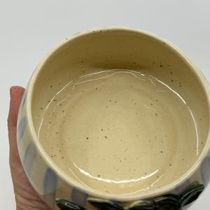 May include: A ceramic bowl with a cream-colored interior and a light blue striped exterior. The bowl is decorated with purple fruit and green leaves. The bowl is round and has a speckled texture.