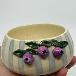 A ceramic bowl with a cream-colored interior and exterior. The exterior features blue vertical stripes and three purple blueberry designs with green leaves. The bowl is held in a hand.