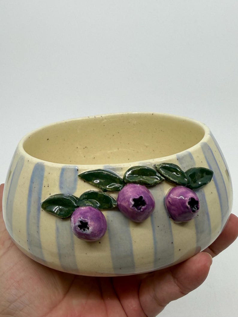 A ceramic bowl with a cream-colored interior and exterior. The exterior features blue vertical stripes and three purple blueberry designs with green leaves. The bowl is held in a hand.