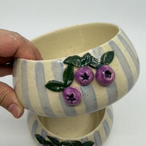 Two ceramic bowls, one atop the other, with a cream-colored interior and blue and cream striped exterior. Each bowl is decorated with three purple blueberries and green leaves. The bowls are handmade and have a rustic, artisanal feel.