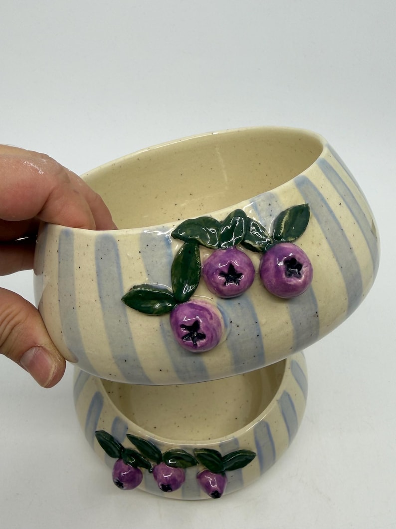 Two ceramic bowls, one atop the other, with a cream-colored interior and blue and cream striped exterior. Each bowl is decorated with three purple blueberries and green leaves. The bowls are handmade and have a rustic, artisanal feel.
