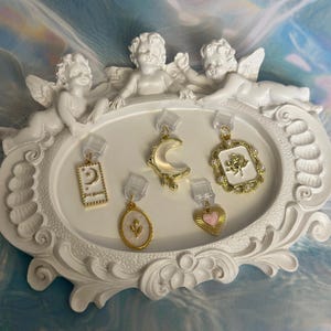 May include: An assortment of gold-coloured earrings featuring moon, heart, and floral motifs, including a rectangular charm with the text "The Moon". The earrings are presented on a white, decorative tray adorned with cherub sculptures.