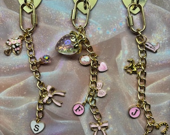 Op de afbeelding: Drie goudkleurige sleutelhangers met hartvormige sluitingen. Elke sleutelhanger heeft een ketting met diverse bedels, waaronder strikken, initialen, een paddenstoel, een aardbei en een hoefijzer. De bedels zijn in roze, goud en transparante kleuren.