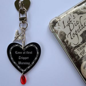 Puede incluir: Un llavero negro en forma de corazón con un borde de alambre de espino blanco y el texto "Love at first Trigger Warning" en fuente gótica. Un pequeño dije de polilla plateado y una cuenta roja translúcida en forma de lágrima cuelgan.