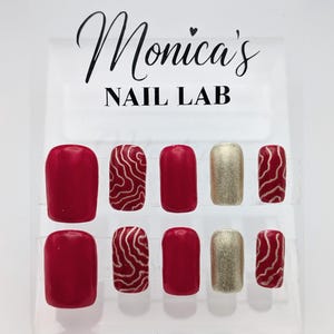 May include: A set of press-on nails in a clear plastic package. The nails are a mix of solid red, gold glitter, and red with gold swirl designs. The package is labelled "Monica's NAIL LAB".