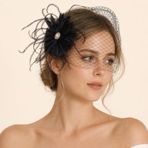 Peut inclure: Une coiffe fascinator noire ornée d'une fleur en plumes sombres avec une perle centrale et des strass. L'accessoire comprend un voile en résille noire fine qui s'étend sur la partie supérieure du visage, créant un aspect structuré.