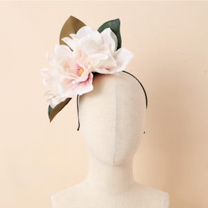 May include: A floral headband with large, white and pink magnolia flowers with yellow centres, and green and brown leaves. The flowers are attached to a thin, black headband, suitable for special events.