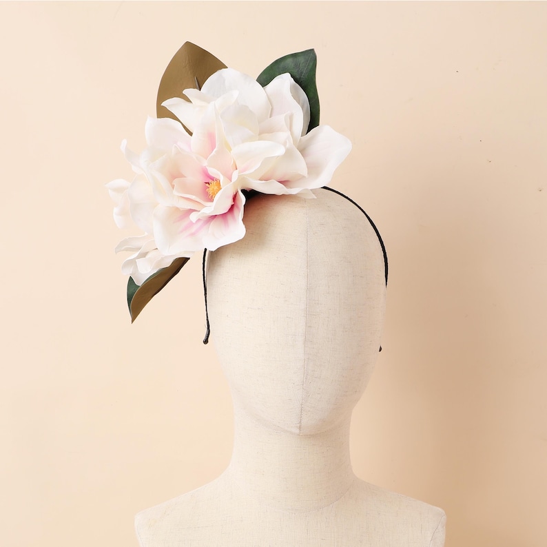 May include: A floral headband with large, white and pink magnolia flowers with yellow centres, and green and brown leaves. The flowers are attached to a thin, black headband, suitable for special events.