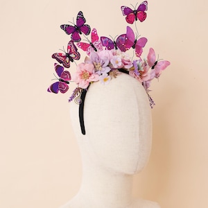 Può includere: Una fascia nera adornata con un gruppo di fiori rosa, viola e lavanda, e diverse decorazioni a farfalla viola e rosa. Le farfalle sono di varie dimensioni e design, aggiungendo un tocco stravagante all'arrangiamento floreale.