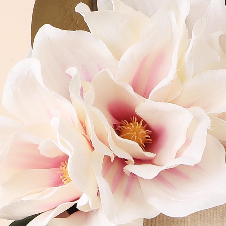 May include: Close-up of artificial magnolia flowers. The petals are a soft white with pink accents near the centre, surrounding a yellow stamen. The flowers are clustered together, creating a full, textured appearance.