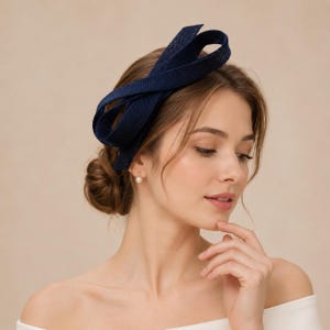 May include: A navy blue fascinator headpiece featuring a structured, looped bow design made from a textured, woven fabric. The accessory is positioned on the side of the head, showcasing a stiff, sculptural form with multiple overlapping loops.