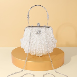 Può includere: Una pochette bianca a forma di conchiglia ricoperta di piccole perle artificiali, dotata di una struttura in metallo argentato, una chiusura superiore con cristallo, un decoro floreale in cristallo sul davanti, un manico curvo in metallo e una sottile catena argentata.