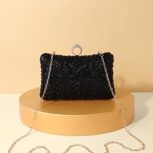 Op de afbeelding: Een rechthoekige zwarte clutch bedekt met ingewikkeld zwart kraalwerk en lovertjes. De tas is voorzien van een goudkleurige metalen kettingriem en een ronde sluiting aan de bovenkant, versierd met heldere kristallen in een goudkleurig frame.
