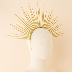 Puede incluir: Una corona dorada con forma de sol, con puntas radiantes de diferentes longitudes. La corona está colocada sobre una fina banda y se muestra sobre una cabeza de maniquí blanca, sobre un fondo neutro. Adecuada para disfraces u ocasiones especiales.