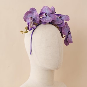 Peut inclure: Une couronne de fleurs d'orchidées violettes. La couronne présente plusieurs grandes fleurs d'orchidées violettes vibrantes fixées sur un fin bandeau violet. Les fleurs ont des nervures détaillées et un aspect réaliste.