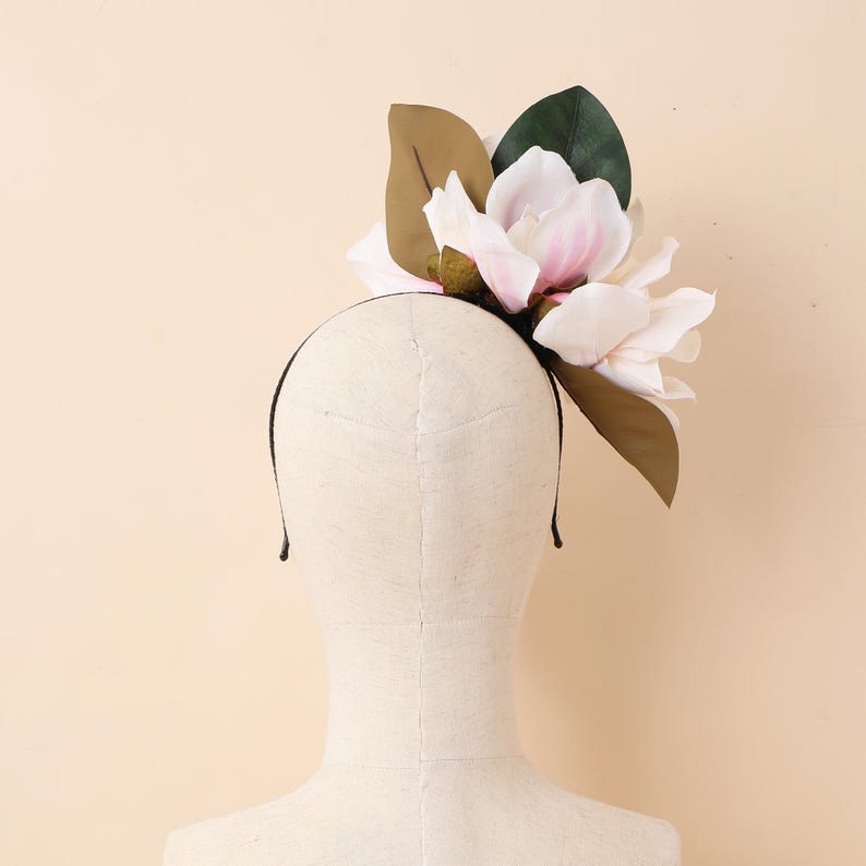 May include: A black headband featuring a cluster of large, lifelike magnolia flowers in white and pink, with green and brown leaves. The floral piece is displayed on a mannequin head.