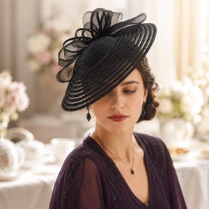 May include: A black fascinator hat featuring a wide, circular, ribbed brim and a large, structured bow made of sheer, layered fabric loops. The hat is positioned at an angle on the head, showcasing a detailed, concentric circular texture.