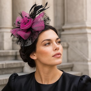 Könnte beinhalten: Ein Fascinator-Kopfschmuck mit leuchtend magentafarbenem Netzstoff in Blütenform, akzentuiert durch schwarze Federn, schwarzes Netz mit Punkten und kleine schwarze Perlen. Das Accessoire ist mit einem dünnen Band am Kopf befestigt.