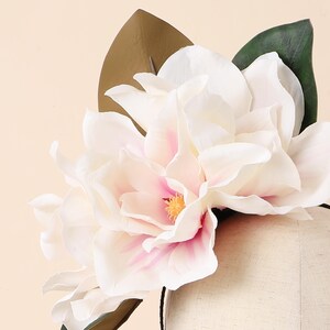 May include: A white and pink magnolia flower with a yellow centre and green and brown leaves. The flower is attached to a black headband, creating a floral headpiece. The petals have a soft, delicate appearance.