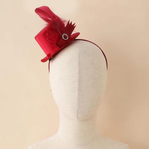 Può includere: Un mini cappello a cilindro rosso su una fascia, ornato da una piuma, un abbellimento scintillante e un ritaglio decorativo. Il cappello è di un rosso vibrante, perfetto per eventi a tema o feste in costume.