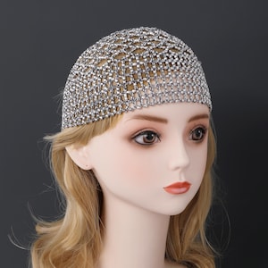 May include: A silver-coloured head covering in a mesh design, densely decorated with small, sparkling rhinestones. The headpiece is displayed on a mannequin with blonde hair and a neutral background, highlighting its intricate design and reflective qualities.
