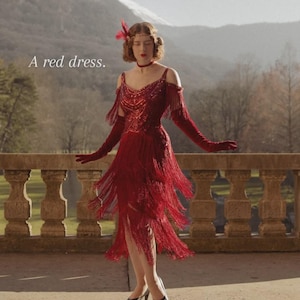 May include: A red dress with a sequined bodice, cold-shoulder sleeves, and multiple tiers of long fringe. The garment features a choker-style neckline and is paired with matching red opera-length gloves and a feathered headpiece. Text reads: A red dress.