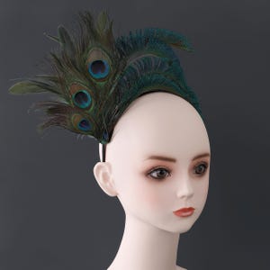 Op de afbeelding: Een decoratieve haarband met pauwenveren in groen, blauwgroen en bruin. De veren zijn in een waaier vorm gerangschikt en bevestigd aan een zwarte haarband. Het accessoire wordt getoond op een mannequinhoofd.