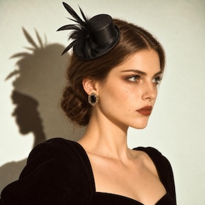 May include: A small black satin mini top hat fascinator adorned with black feathers, paired with a gold-toned rectangular earring featuring a dark central stone. The hat is positioned on the side of the head, showcasing a smooth fabric finish.