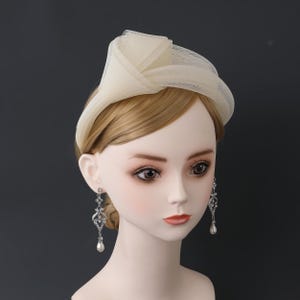 May include: A cream-coloured fascinator with a netted overlay, worn on a mannequin. The headpiece is paired with silver-toned drop earrings with clear stones and a pear-shaped pearl. The mannequin has blonde hair and a neutral background.