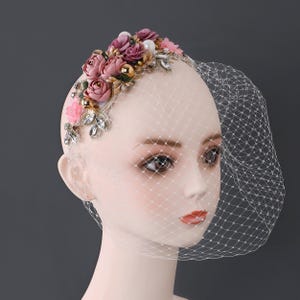 Puede incluir: Un tocado con rosas de tela malva, pequeñas flores rosas, esferas metálicas doradas y cristales transparentes facetados sobre una base decorativa. Un velo tipo jaula blanco con un patrón de malla en forma de diamante está unido al tocado.