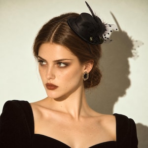 Peut inclure: Un mini chapeau haut-de-forme noir de type bibi avec un petit bord, une plume foncée et un voile blanc à pois noirs. L'accessoire est porté de côté et associé à un vêtement noir ainsi qu'à une boucle d'oreille dorée rectangulaire à pierre noire.