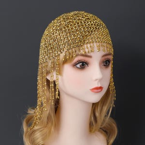 May include: A gold-colored headpiece with a net-like design, adorned with dangling beaded strands. The head covering is made of small, shiny beads, creating a shimmering effect. The headpiece is designed to cover the top of the head and cascade down the sides.