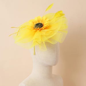 May include: A vibrant yellow headpiece with a sunflower and tulle detailing. The sunflower has a dark centre and is surrounded by yellow feathers. The headpiece is designed to be worn on the head.