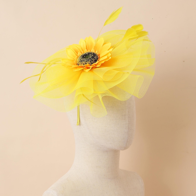 May include: A vibrant yellow headpiece with a sunflower and tulle detailing. The sunflower has a dark centre and is surrounded by yellow feathers. The headpiece is designed to be worn on the head.