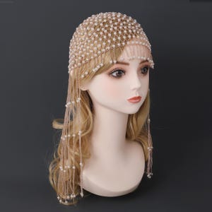 Könnte beinhalten: Ein beiges Kopfstück mit netzartigem Design, verziert mit kleinen, runden, weißen Perlen. Das Kopfstück hat einen Perlenbesatz, der herunterhängt. Das Kopfstück wird auf einem Schaufensterkopf mit blonden Haaren präsentiert.