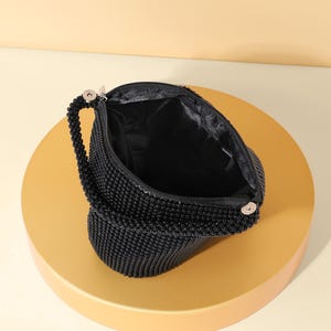 Op de afbeelding: Een zwarte kralentas met een textuur van kleine, uniforme zwarte kralen. De tas heeft een enkel kralenhandvat bevestigd met zilverkleurige metalen hardware en een met zwarte stof gevoerde binnenkant met een ritssluiting.