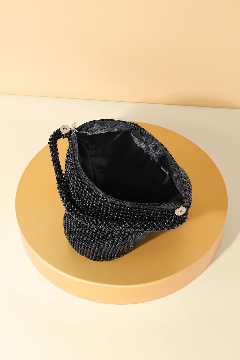 Op de afbeelding: Een zwarte kralentas met een textuur van kleine, uniforme zwarte kralen. De tas heeft een enkel kralenhandvat bevestigd met zilverkleurige metalen hardware en een met zwarte stof gevoerde binnenkant met een ritssluiting.