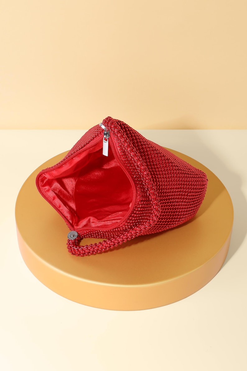 Op de afbeelding: Een felrode handtas bedekt met kleine, uniforme kralen met een getextureerd oppervlak. De tas heeft een enkel kralenhandvat en een zilverkleurige metalen ritssluiting aan de bovenkant. De binnenkant is gevoerd met bijpassende rode stof, zichtbaar door de opening.