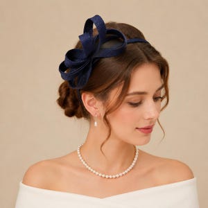 May include: A navy blue fascinator headband featuring a large, sculptural bow made of stiffened, woven fabric loops. The accessory is positioned on the side of the head, showcasing a textured, dark blue material with a structured, layered design.