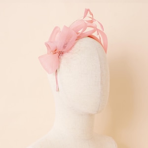 May include: A pale pink headband featuring a bow and delicate feather detailing. The headband is constructed from a slender, pliable material and includes a prominent bow on the left. The feathers are arranged in a fan, providing a refined aesthetic.