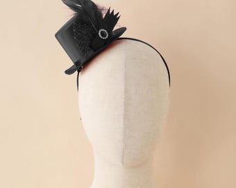May include: A black mini top hat headband with a feather and jewel embellishment. The hat features a small, structured design with a curved brim. A black headband secures the hat to the head. The hat is decorated with a black feather and a sparkling jewel.