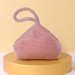Op de afbeelding: Een ros&eacute;gouden handtas bedekt met kleine, reflecterende metalen kralen. De tas heeft een driehoekig, soepel silhouet met een enkele ge&iuml;ntegreerde ronde handgreep aan de bovenkant, die volledig is versierd met dezelfde ros&eacute;gouden kralen.