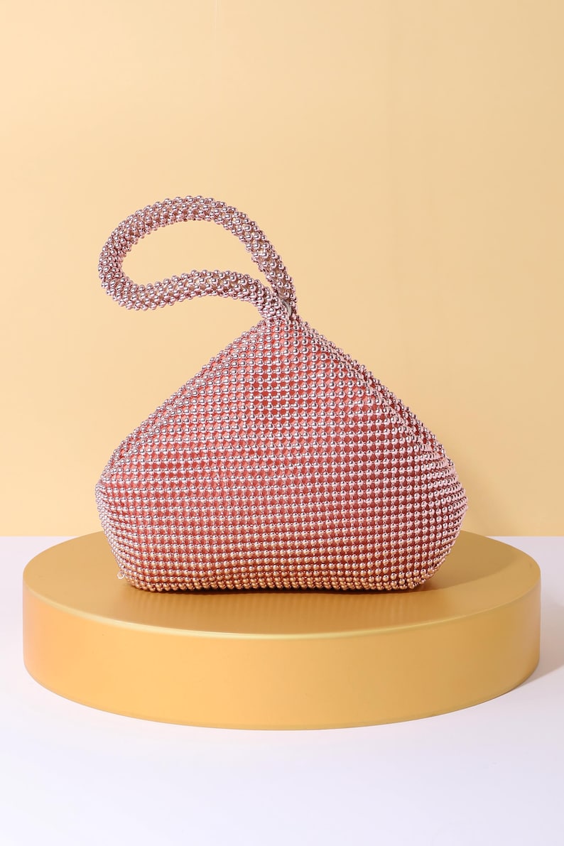 Op de afbeelding: Een ros&eacute;gouden handtas bedekt met kleine, reflecterende metalen kralen. De tas heeft een driehoekig, soepel silhouet met een enkele ge&iuml;ntegreerde ronde handgreep aan de bovenkant, die volledig is versierd met dezelfde ros&eacute;gouden kralen.