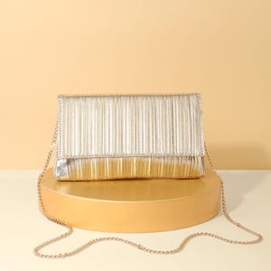 Op de afbeelding: Een rechthoekige metallic gouden clutch met een geplooide textuur op de voorflap. De tas is voorzien van een dunne, delicate schouderriem van goudkleurige metalen ketting die aan de zijkanten van de gestructureerde, rechthoekige tas hangt.