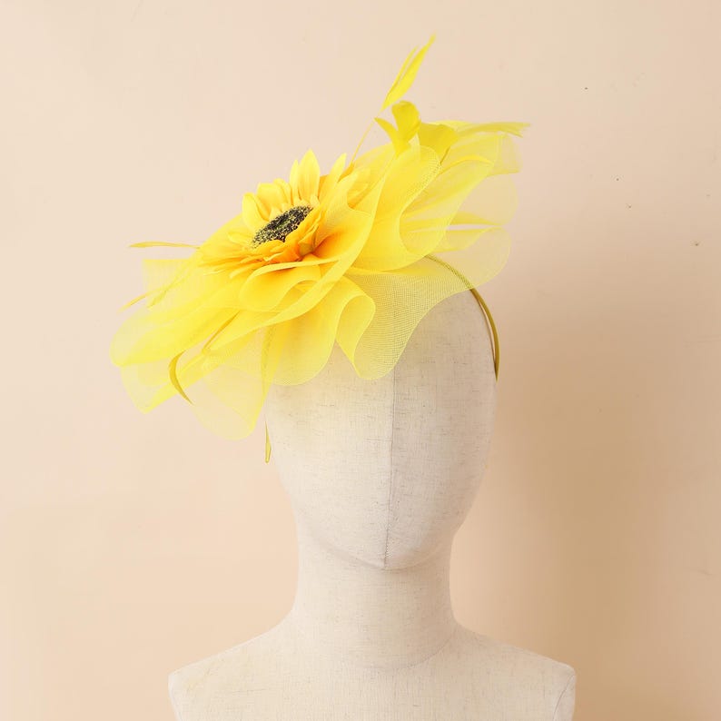 May include: A vibrant yellow sunflower fascinator with a black centre and fine yellow feathers. The headpiece is attached to a thin, yellow headband. The flower is made of layered fabric, creating a voluminous effect. Ideal for special events and celebrations.